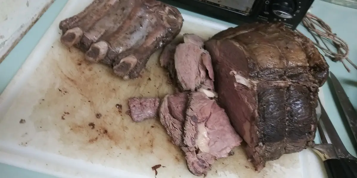 A white cutting board holds a large, sliced roast beef with a dark crust and pink interior, alongside a rack of cooked ribs…