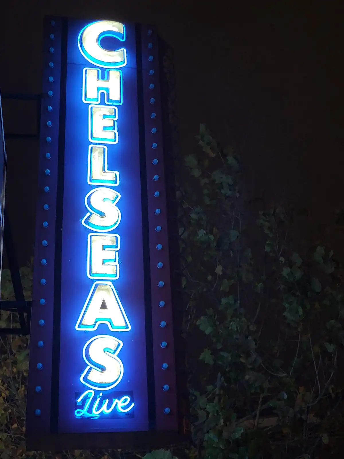 A vertical neon sign at night reads 'CHELSEA'S Live' in glowing blue and white letters, framed by riveted metal sides, with…