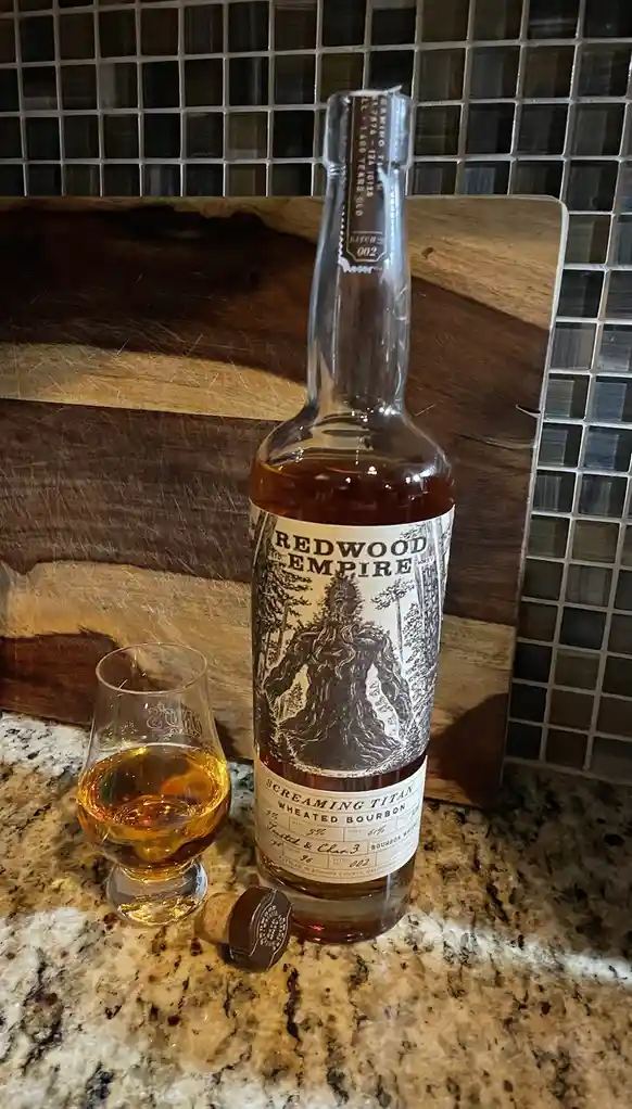 A bottle of Redwood Empire 'Screaming Titan' wheated bourbon sits on a granite countertop next to a tasting glass filled…