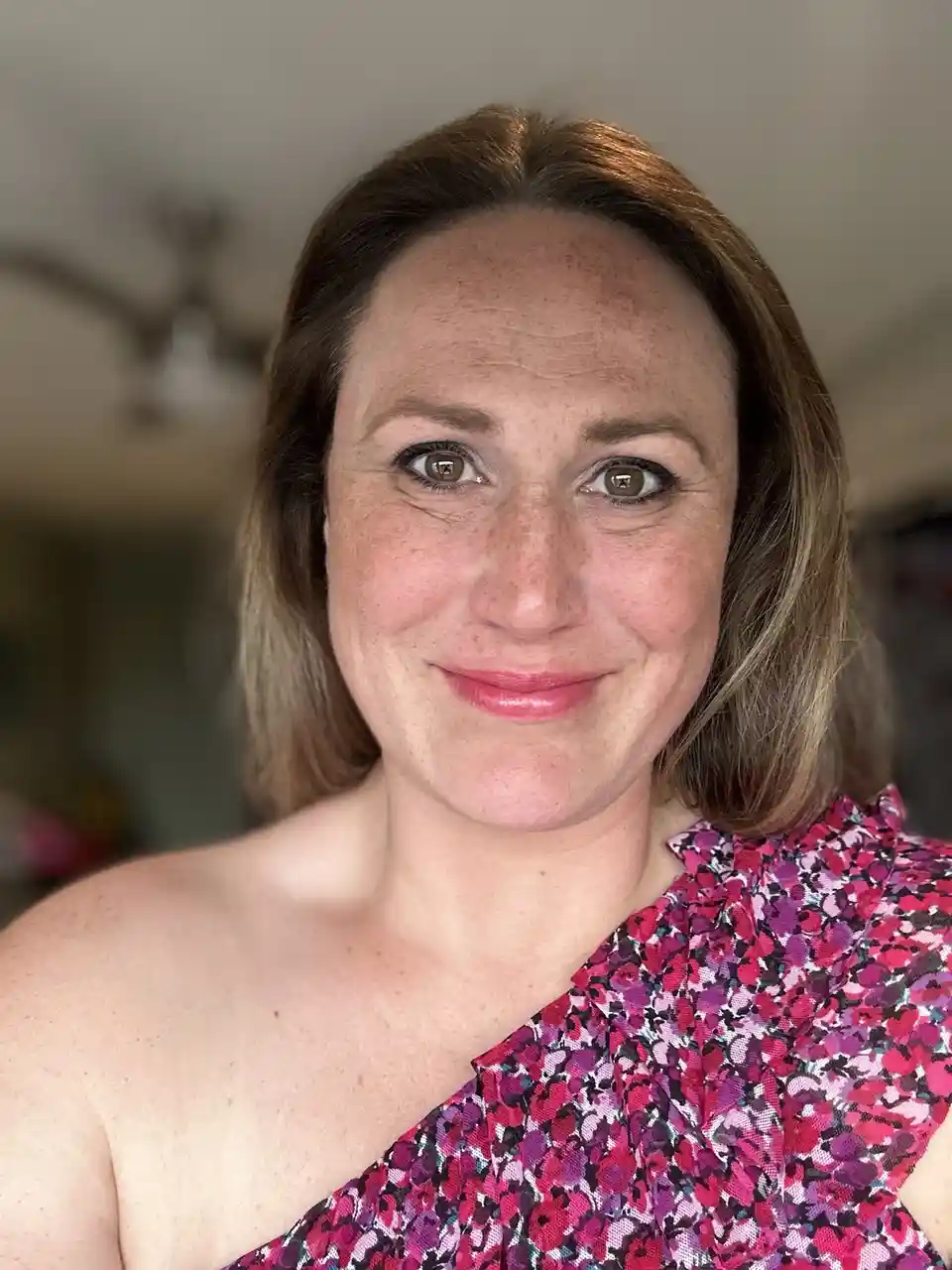 A woman with shoulder-length brown hair and freckles smiles at the camera while wearing a colorful floral one-shoulder top…