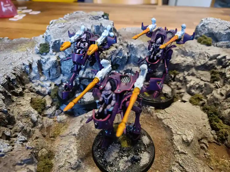Three purple and orange painted sci-fi miniatures stand on rocky terrain, each holding weapons and looking forward. The…