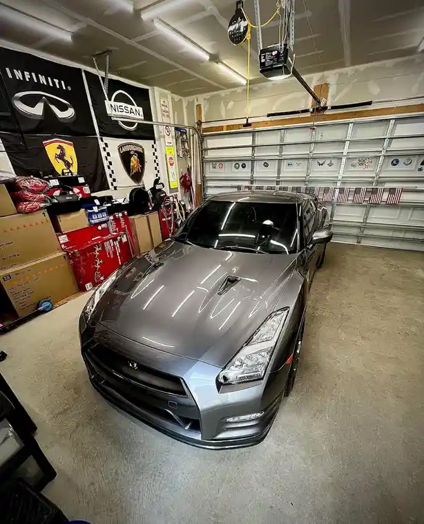 A shiny gray Nissan GT-R is parked in a garage decorated with Infiniti, Nissan, Ferrari, and Lamborghini banners. The car's…