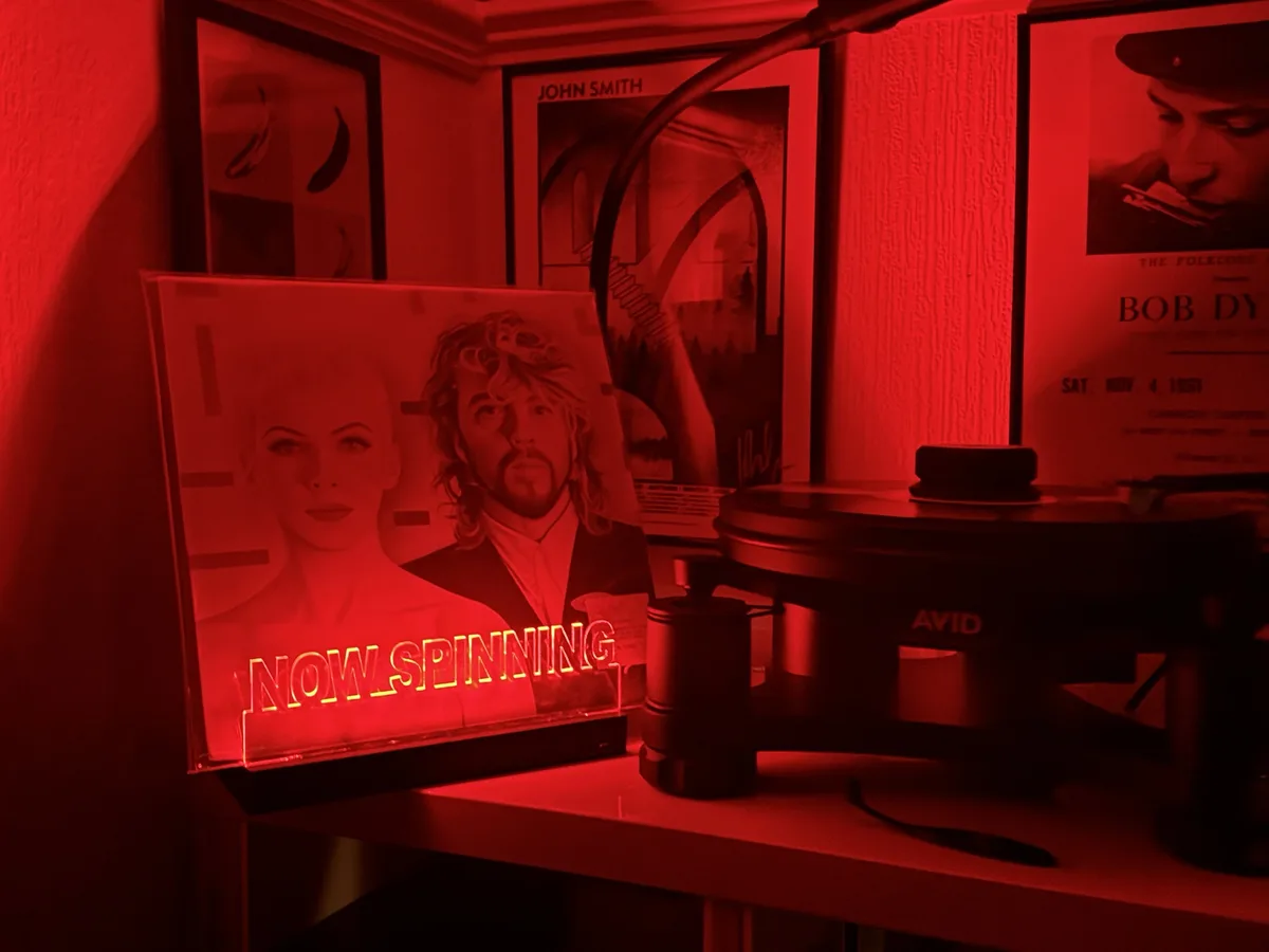 A red-lit room features a turntable with a glowing neon sign reading 'NOW SPINNING' in front of it, displaying images of a…