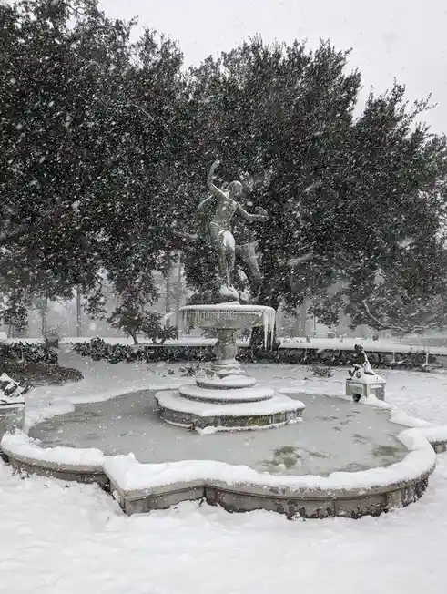 A snow-covered outdoor fountain with a central bronze statue of a figure, surrounded by snow-laden trees and a frozen basin…