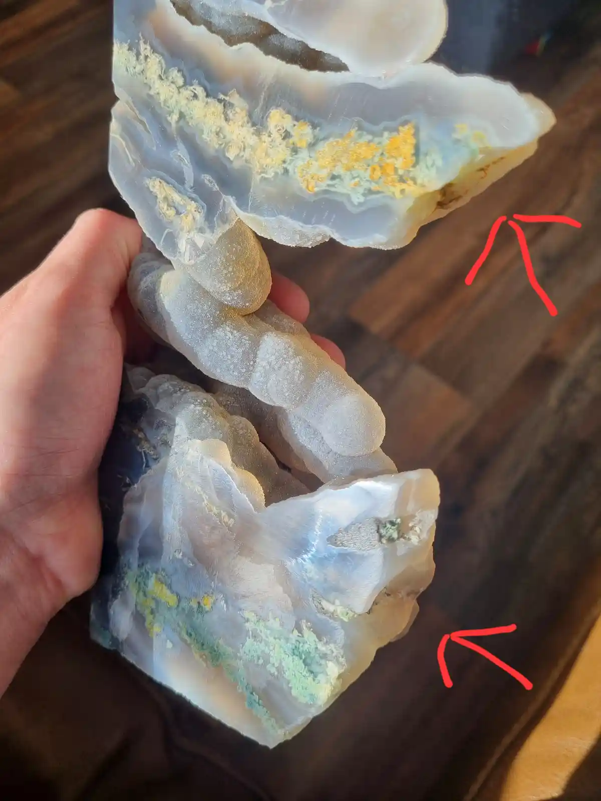 A hand holds a large, irregularly shaped geode with translucent white and blue layers, accented by yellow and green mineral…
