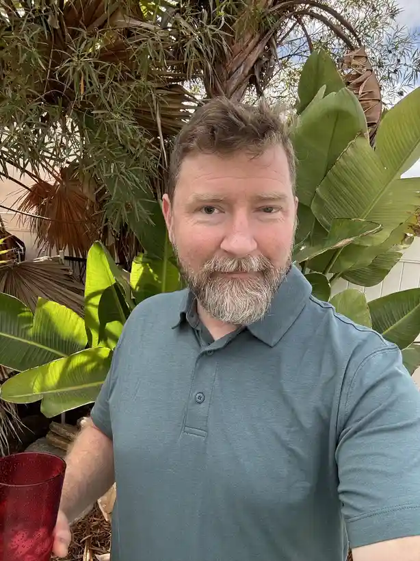 A middle-aged man with a graying beard and short brown hair, wearing a teal polo shirt, holds a red textured glass while…