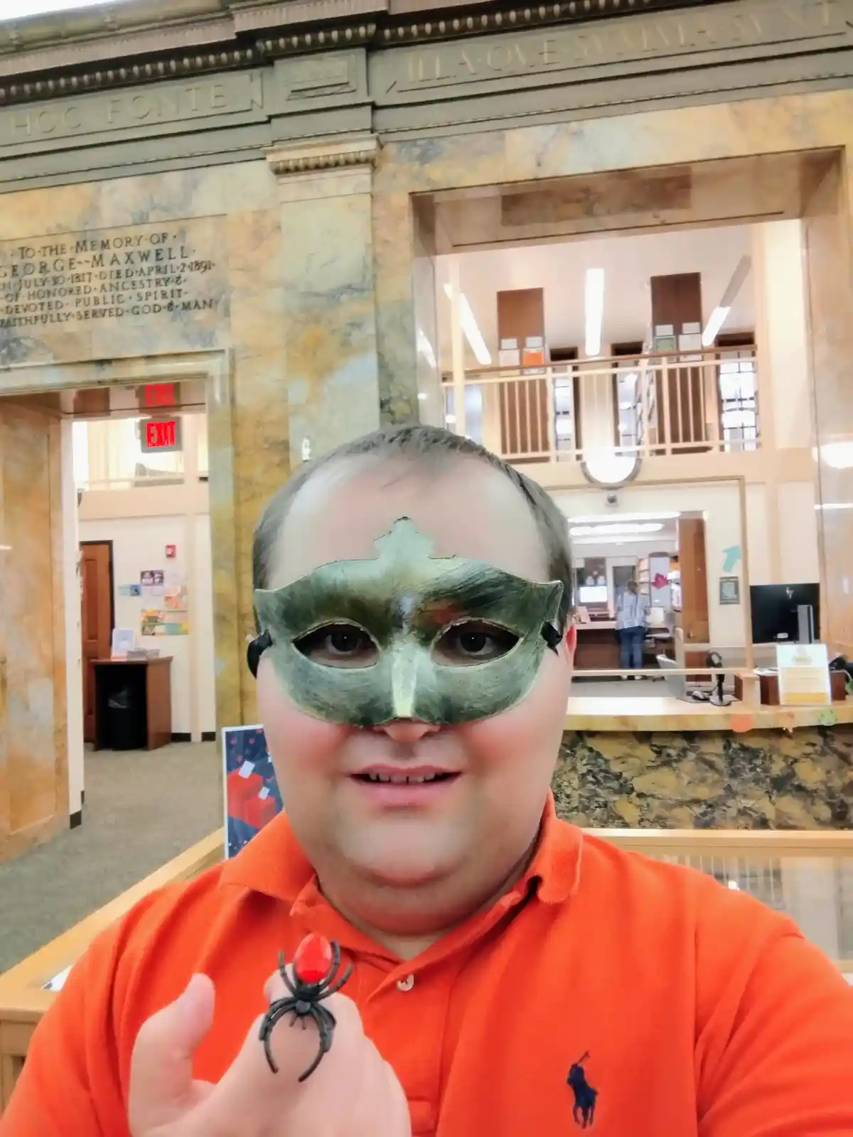 A man wearing a green Venetian-style mask and an orange polo shirt stands inside a grand library with marble walls and an…