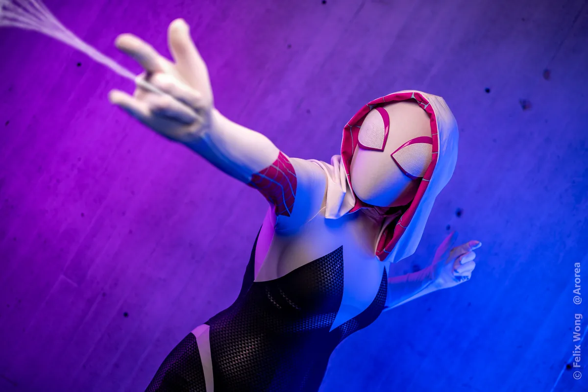 A person in a Spider-Gwen cosplay, wearing a white, black, and pink suit with a hood and web-patterned mask, is posed…