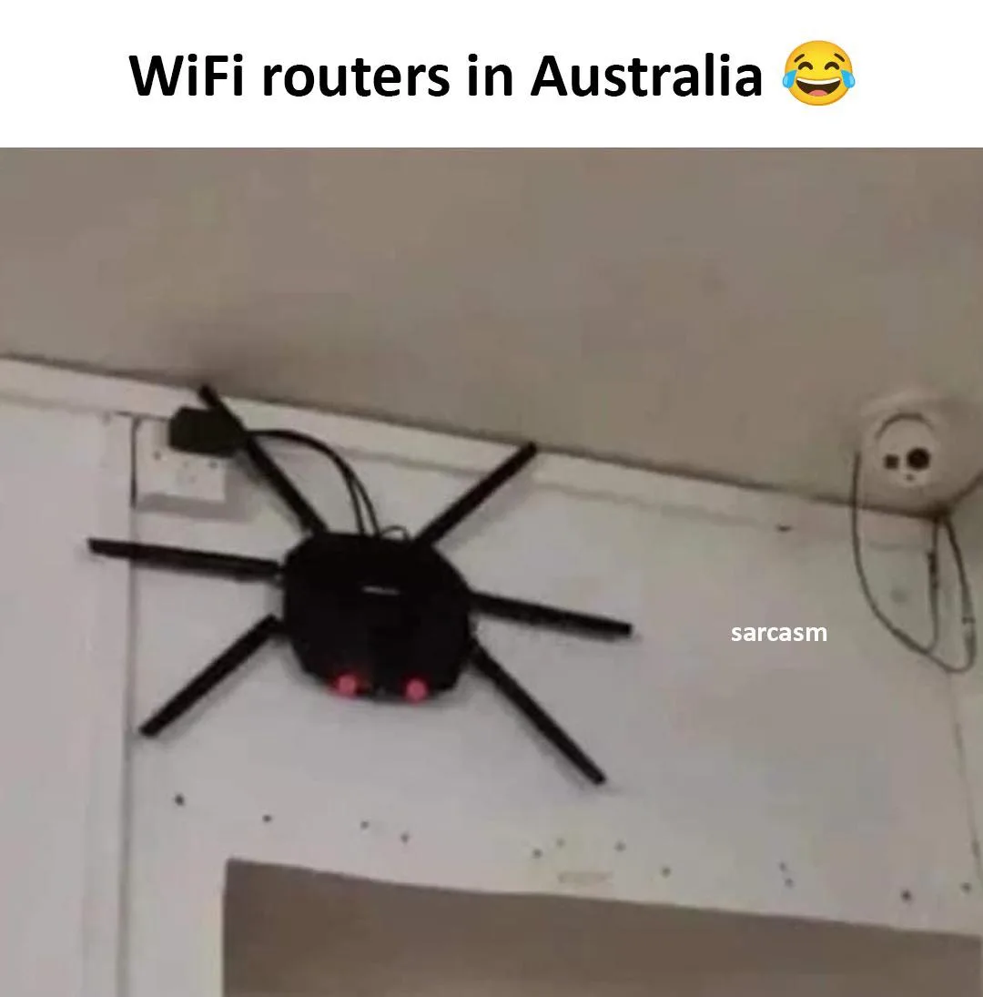 A black WiFi router with multiple antennas is mounted on a white wall, resembling a spider with glowing red lights for eyes…