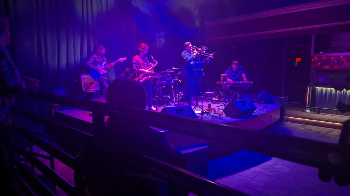 A live band performs on a dimly lit stage bathed in purple and blue lights, featuring a guitarist, saxophonist, trombonist…