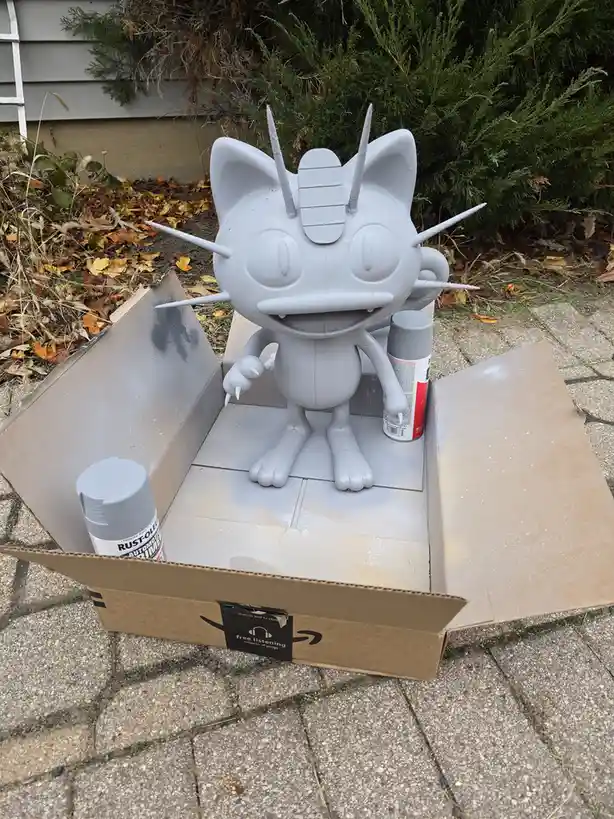 A large, unpainted gray statue of the Pokémon Meowth sits inside an open Amazon cardboard box on a paved patio, surrounded…