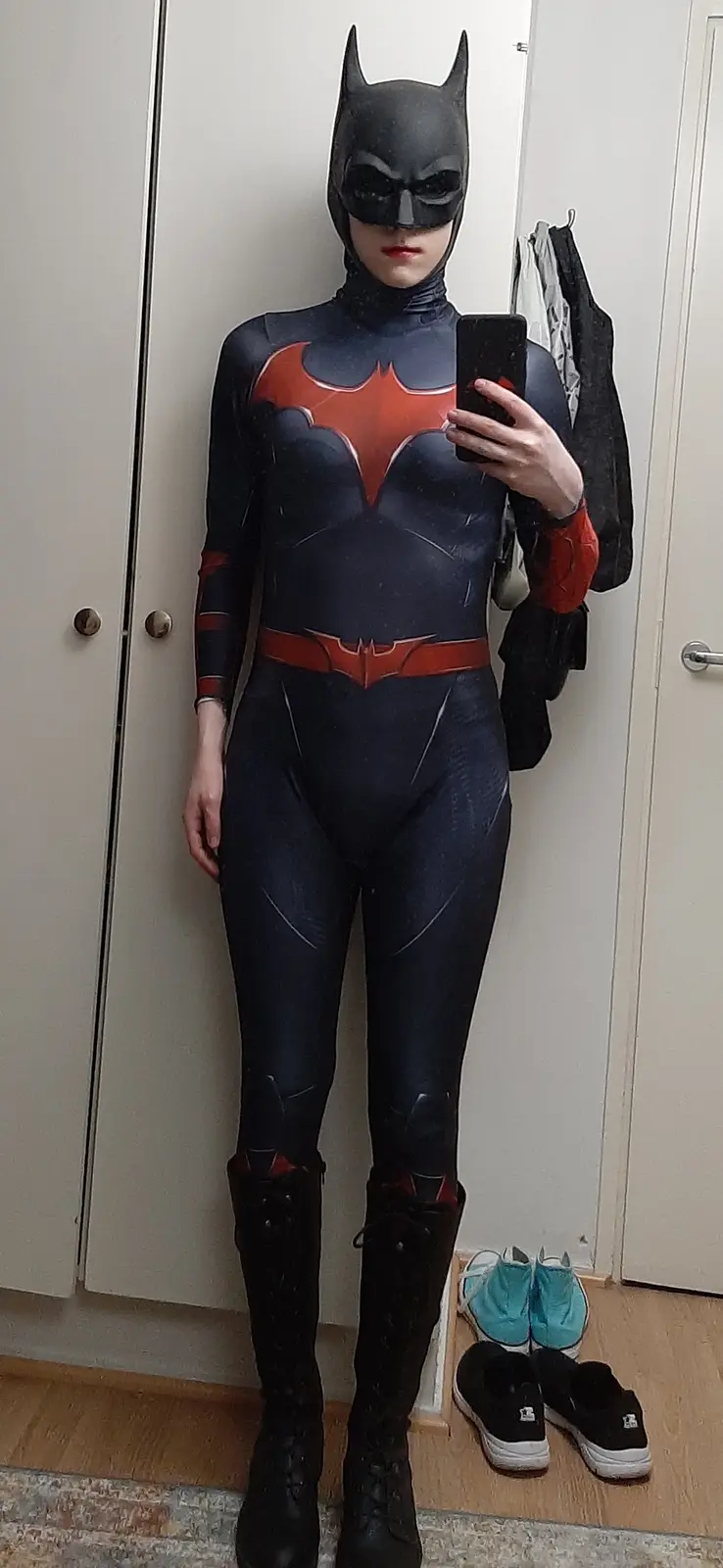 A person in a full-body Batwoman cosplay, featuring a dark blue suit with a red bat emblem on the chest and a matching cowl…