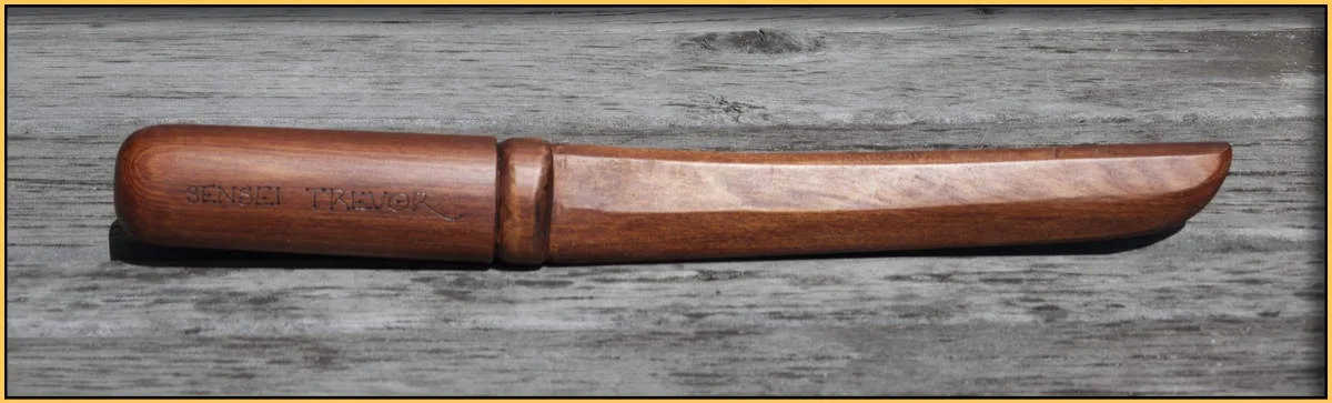 A wooden knife with a handle and blade, both carved from wood, lies horizontally on a weathered gray wooden surface. The…