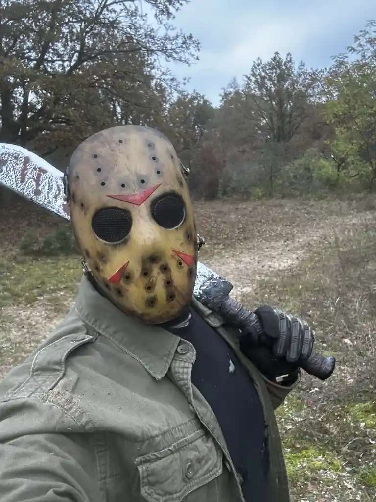 A person wearing a Jason Voorhees hockey mask with red triangular markings, holding a large knife over their shoulder…