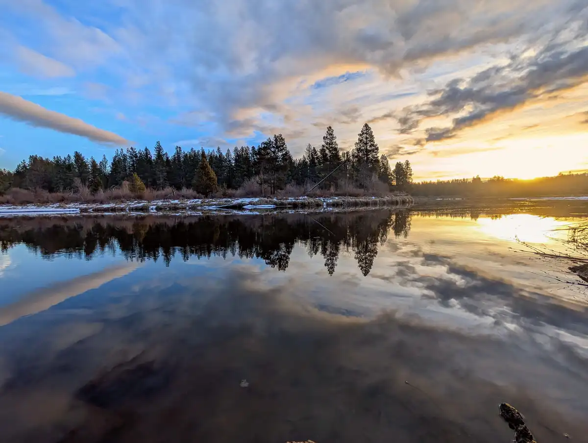 A serene landscape photo shows a calm body of water reflecting a dramatic sunset sky with clouds and distant pine trees. The…