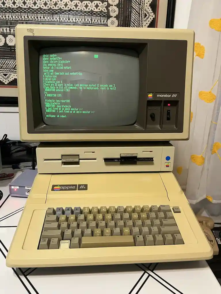 A vintage beige Apple IIc computer with an integrated monitor displays a green text-based chat log. The chat shows users…