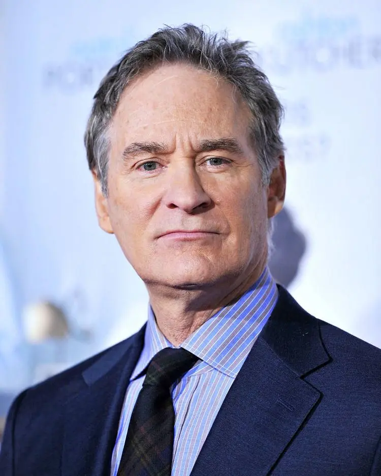A close-up photo of actor Kevin Kline, who has gray hair, is wearing a dark suit, a striped blue and white shirt, and a dark…