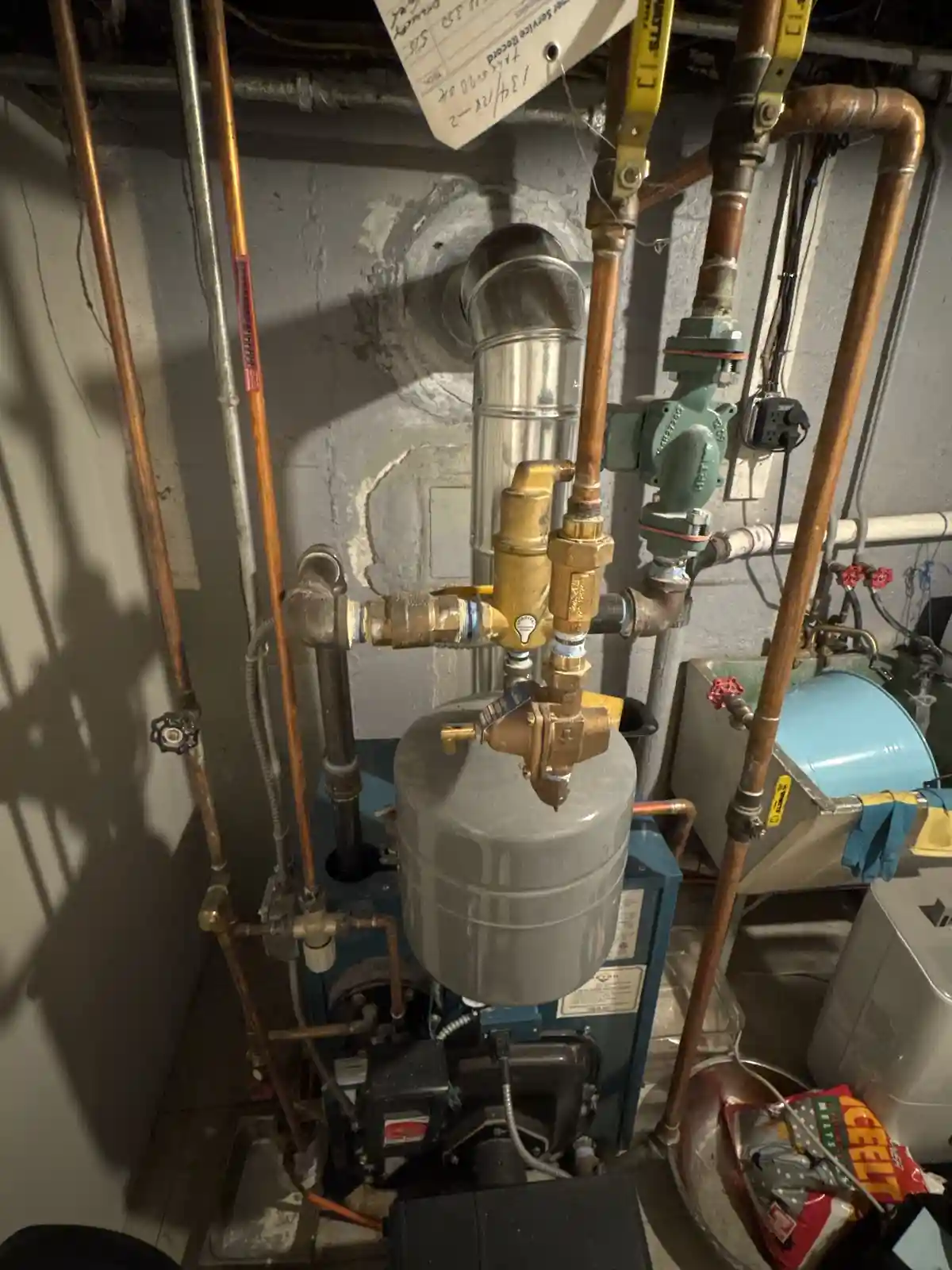 A close-up photo of a complex, cluttered basement heating system with copper pipes, valves, and a gray expansion tank. A bag…