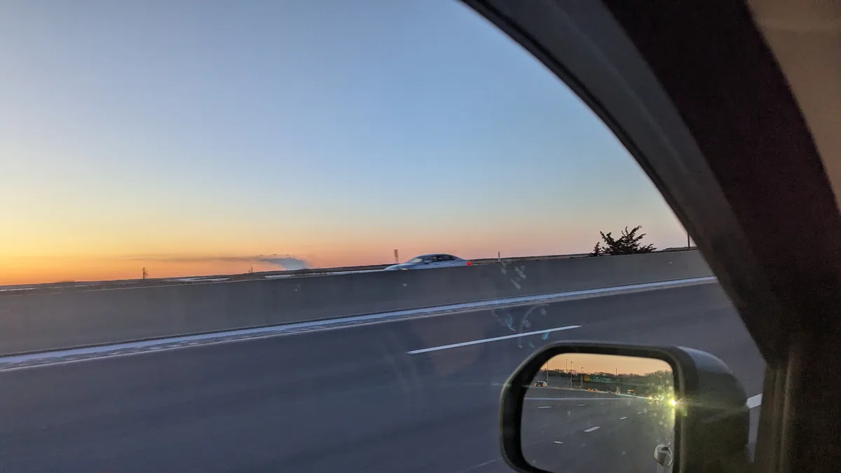 A photo taken from inside a car looking out at a highway during sunset, showing a silver car driving on the opposite side of…