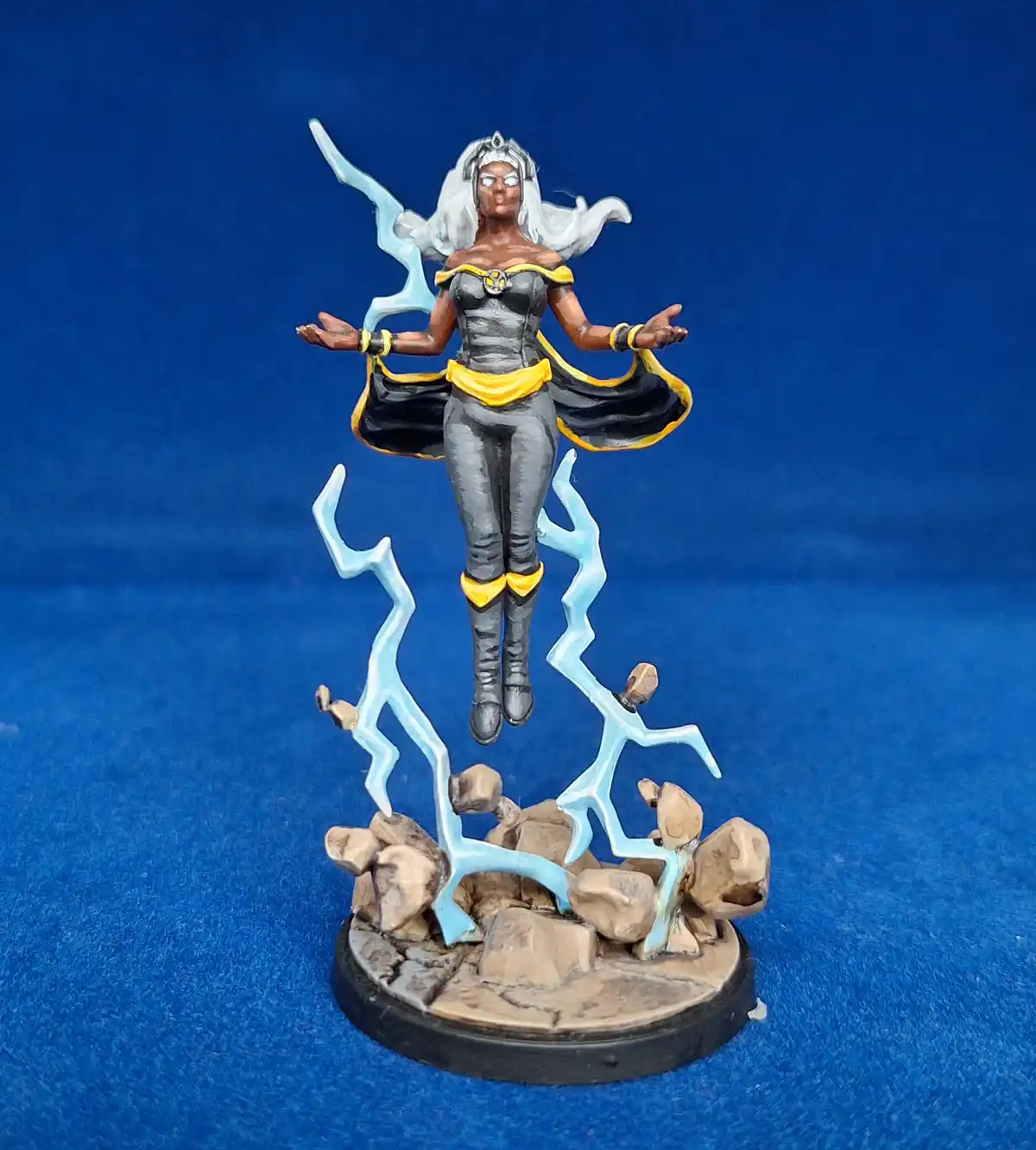 A painted miniature figurine of a female character with long white hair, wearing a black and yellow outfit, levitating above…