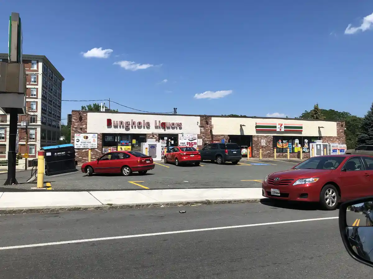 A daytime photo of a strip mall with a 7-Eleven and a liquor store named 'Bunghole Liquors' side-by-side, with several cars…