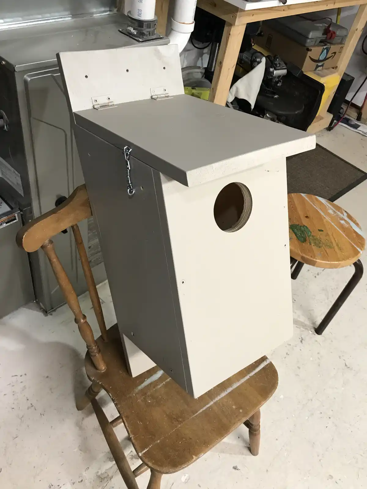 A handmade birdhouse with a grey top and white sides, featuring a round entrance hole and a hinged lid secured with a small…