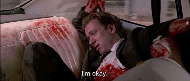 A man is slumped over in a car seat covered in blood, his clothes and arm also stained with blood, looking dazed and…