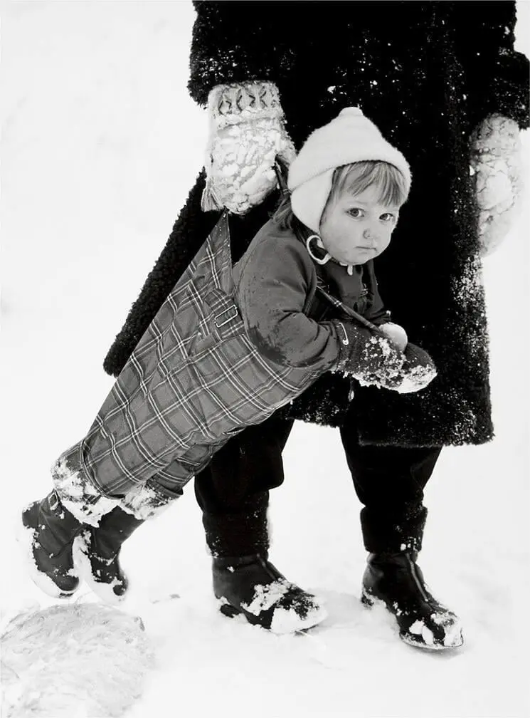 A black-and-white photo shows a young child in a plaid snowsuit and knit hat being carried by an adult in a dark coat and…