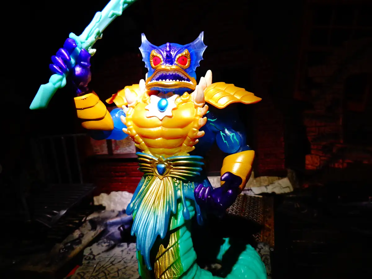 A brightly colored, muscular action figure with a blue fish-like head, yellow torso, and teal lower body, holding a glowing…