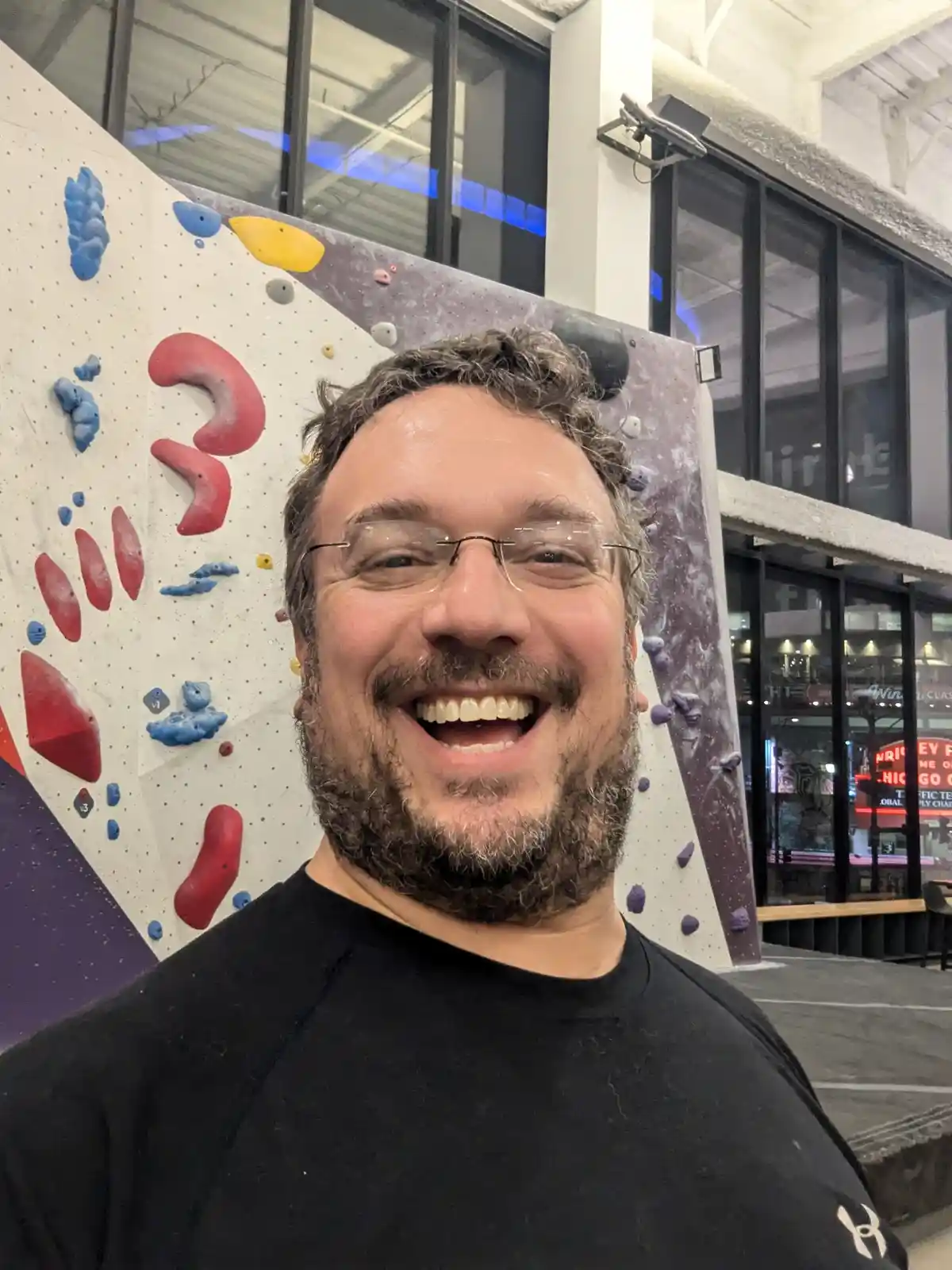 A man with glasses and a beard, wearing a black shirt, smiles broadly at the camera while standing in front of an indoor…