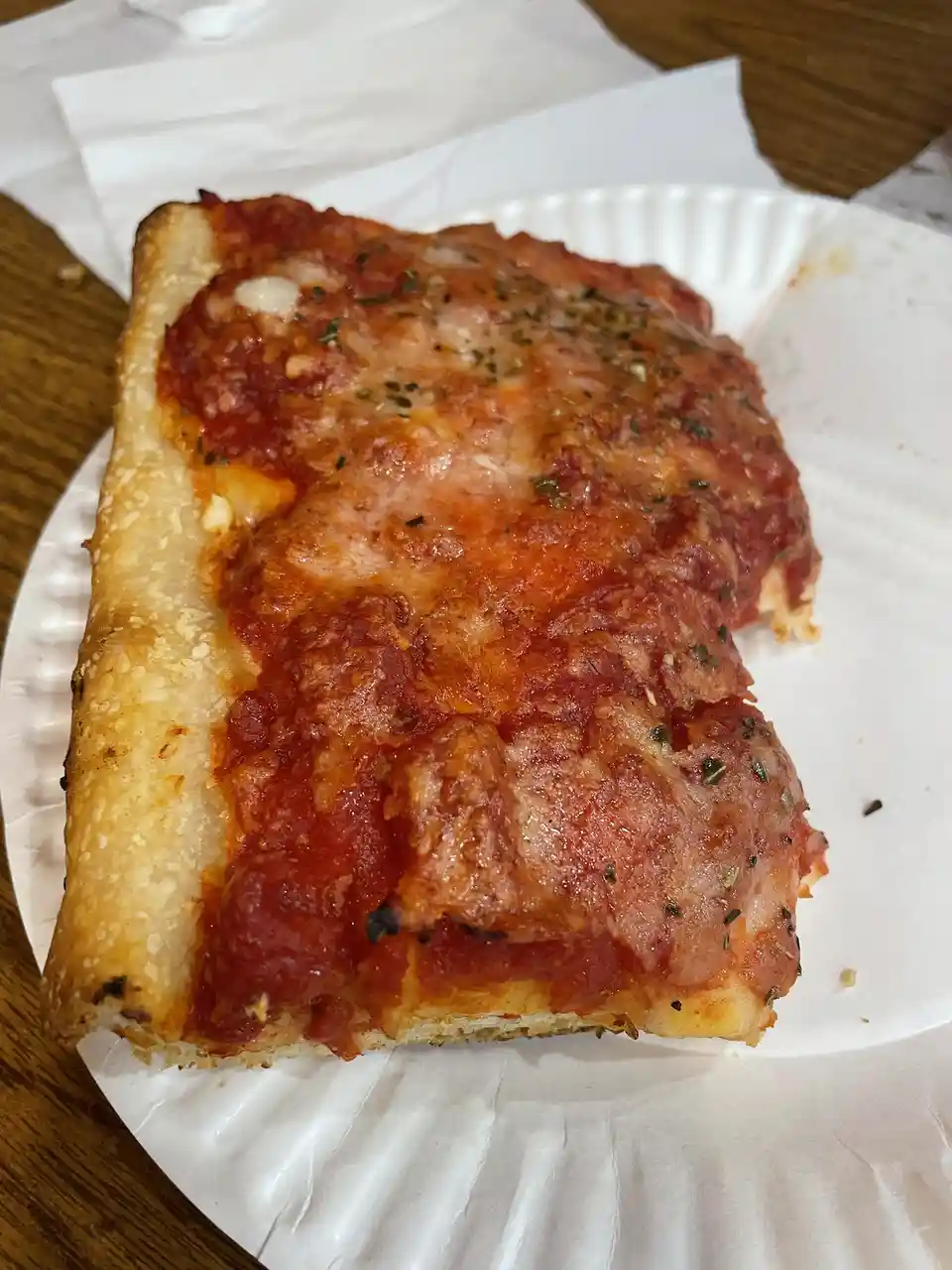 A close-up photo shows a thick, rectangular slice of deep-dish pizza on a white paper plate, resting on a wooden table. The…