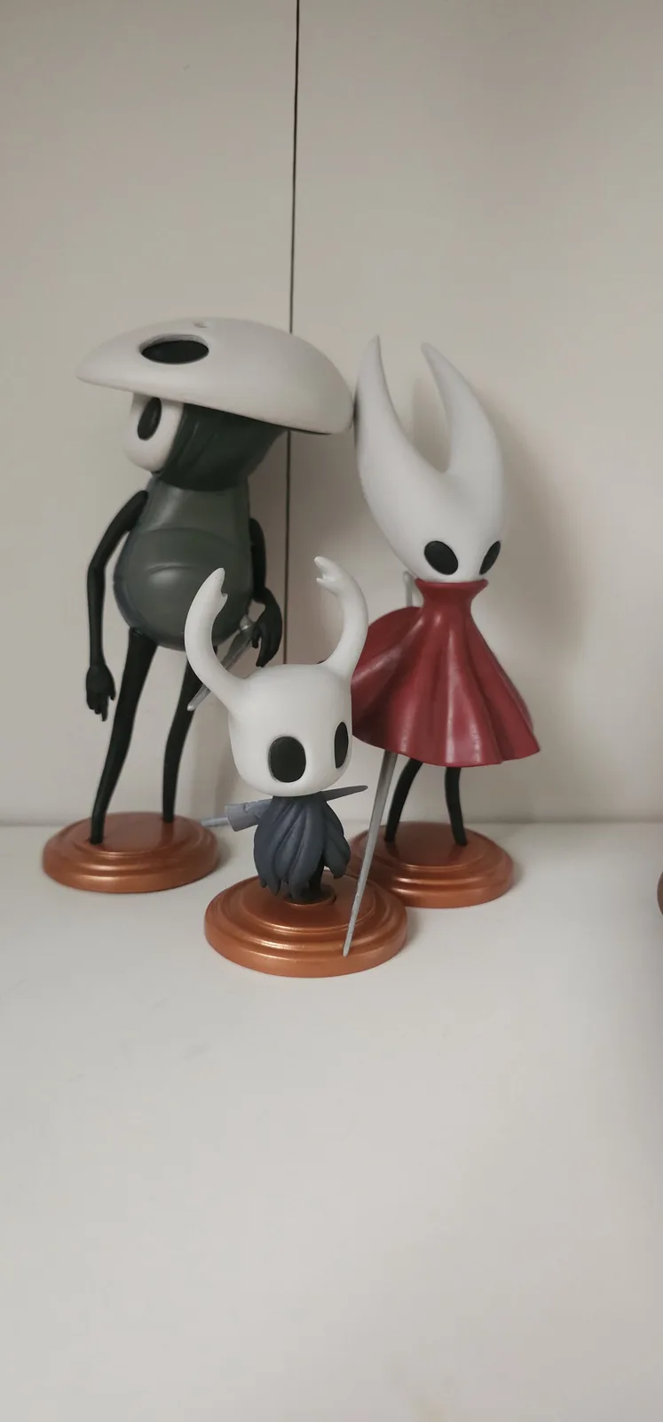 Three Hollow Knight figurines stand on a shelf: a tall grey one with a mushroom cap, a tall white one in a red dress, and a…
