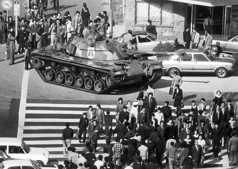A black-and-white historical photograph shows a military tank driving through a busy urban street, with civilians, including…