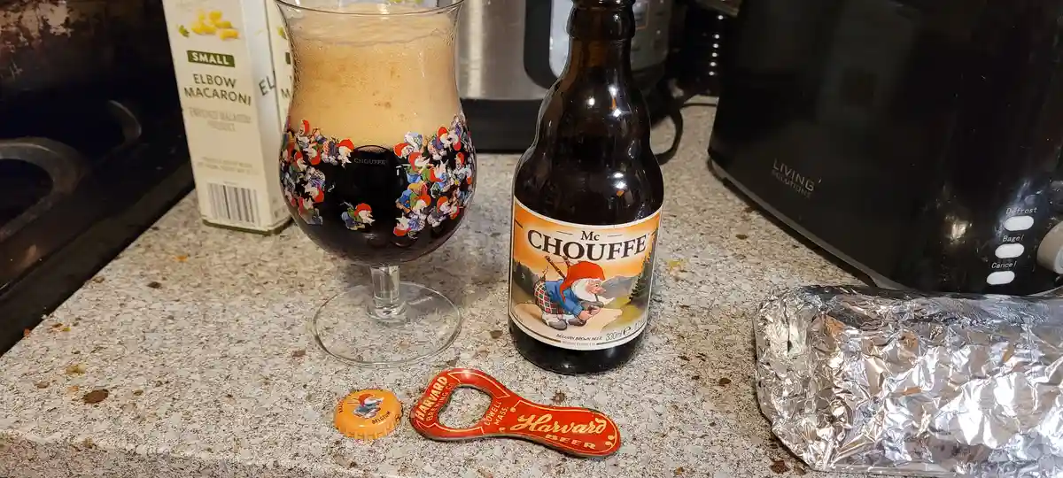A kitchen counter displays a bottle of Mc Chouffe Belgian brown beer next to a glass filled with the dark beer and a thick…