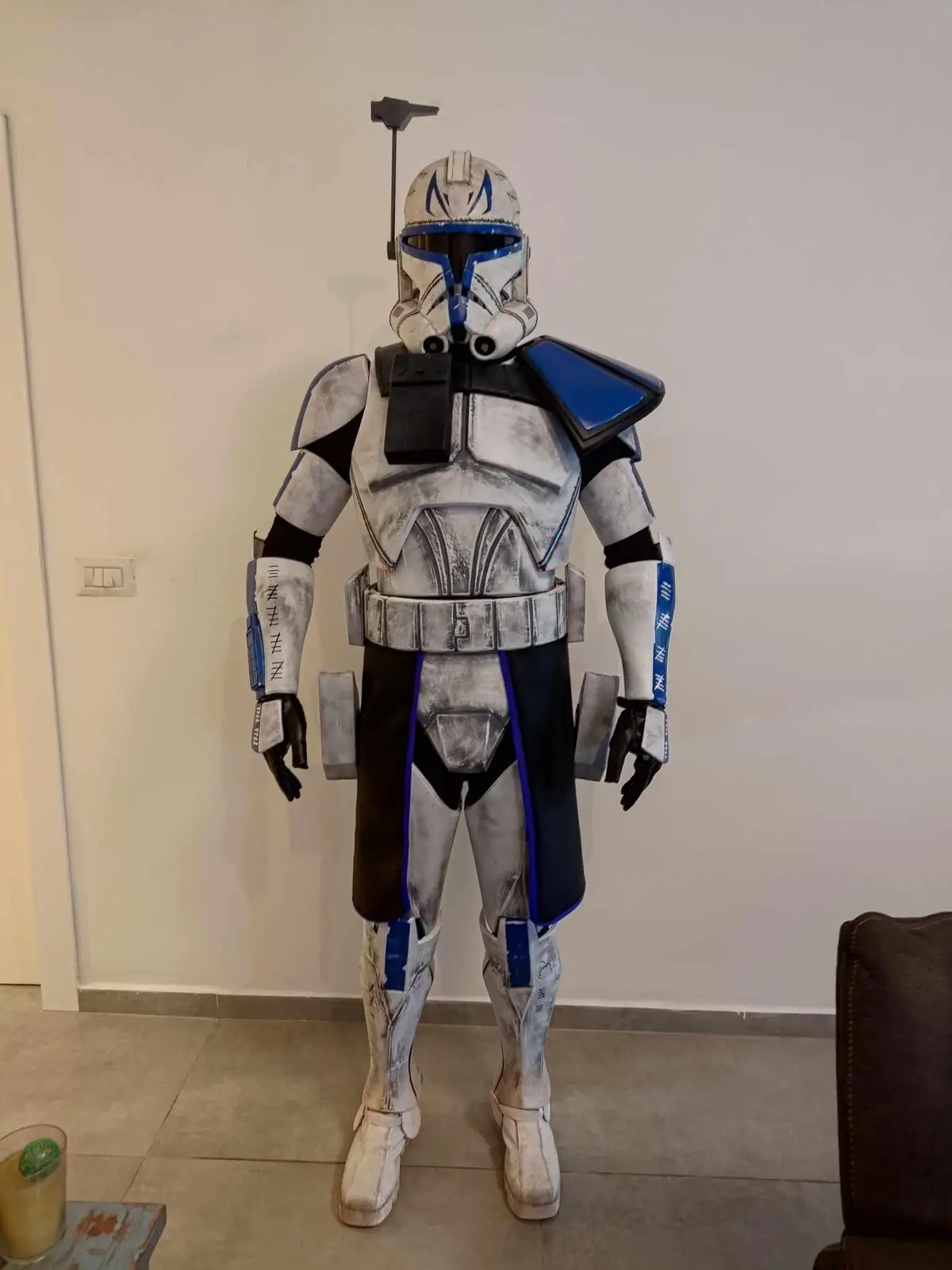 A person in a detailed, weathered Star Wars clone trooper costume with blue and white armor stands indoors against a plain…