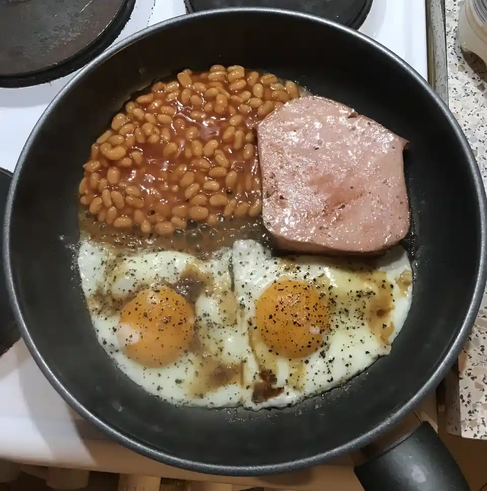 A black frying pan on a stovetop contains a meal with two fried eggs at the bottom, their yolks forming eyes, a slice of…