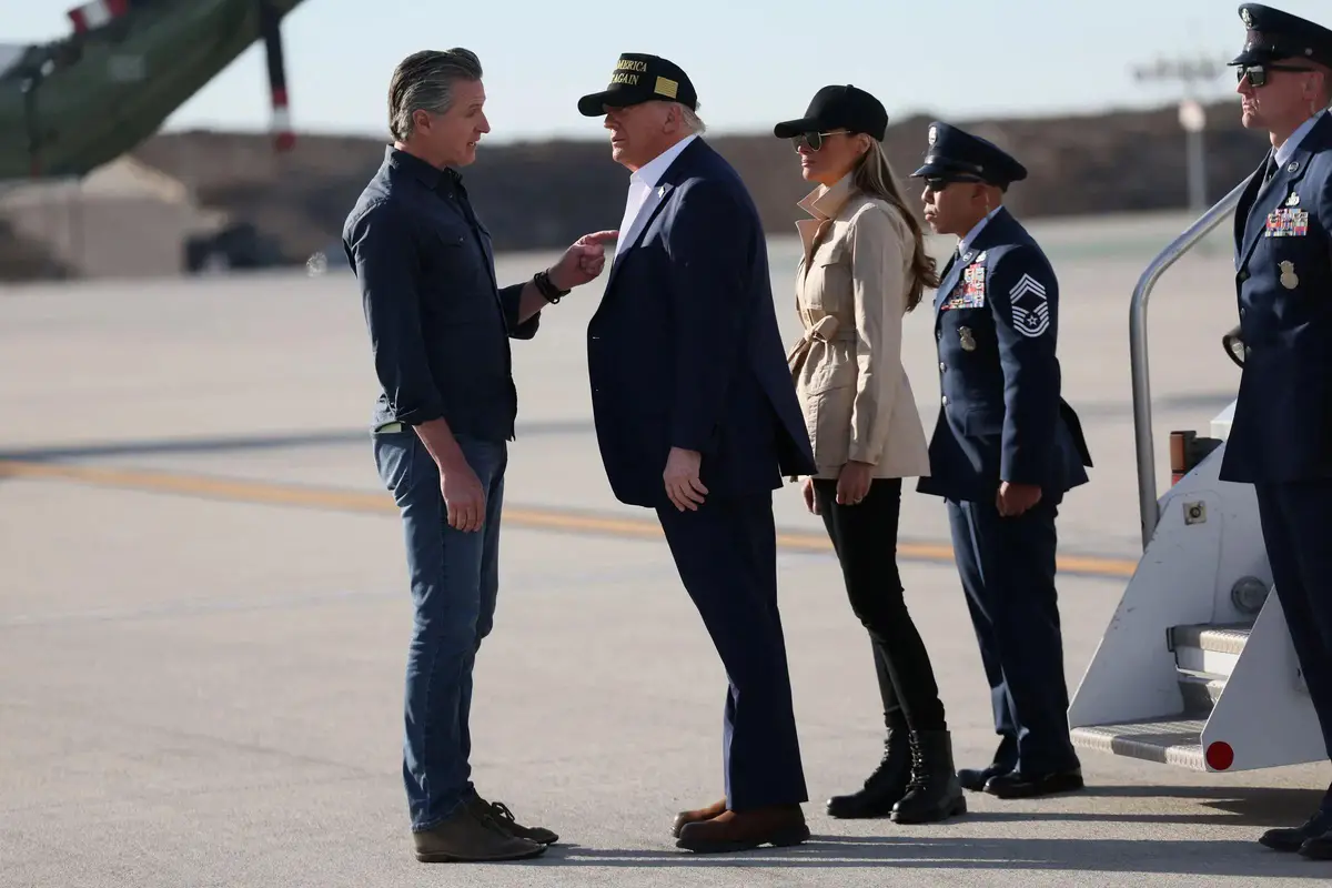 A man in a dark shirt and jeans points at another man in a suit and 'Make America Great Again' cap, who is standing next to…