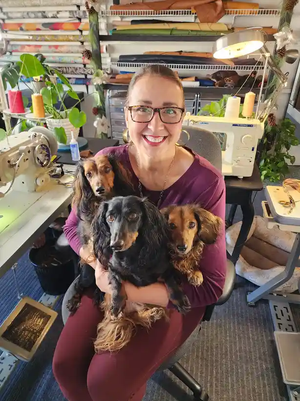 A smiling woman wearing glasses and a purple top sits in a chair in a craft room filled with sewing machines, fabric rolls…