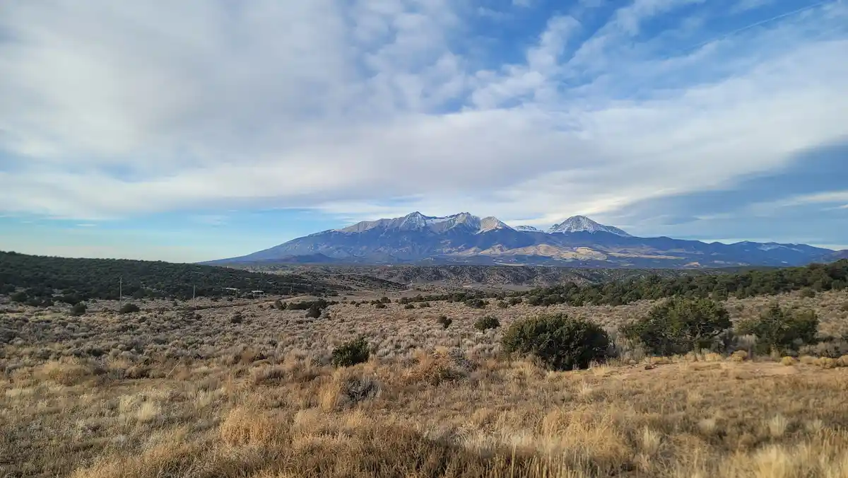 A wide landscape photo shows a dry, grassy plain with scattered shrubs in the foreground, rolling hills in the midground…