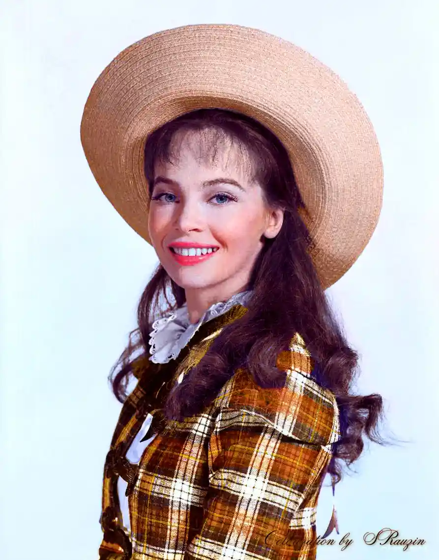 A vintage studio portrait of a smiling woman with long brown hair, wearing a wide-brimmed straw hat and a plaid jacket over…