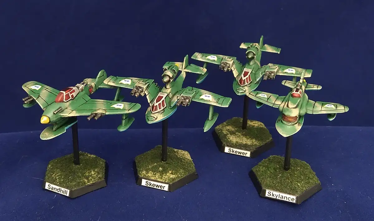 Four green-painted, stylized aircraft miniatures are displayed on hexagonal bases with grass texture against a blue…