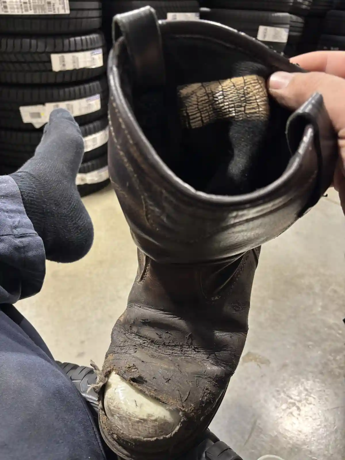 A close-up photo shows a person holding up a very worn, dirty, and torn brown work boot with a visibly damaged toe cap…