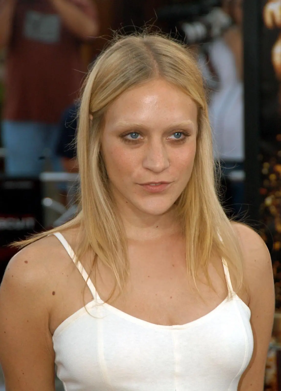 A close-up photo of a blonde woman with blue eyes and a neutral expression, wearing a white tank top, looking directly at…