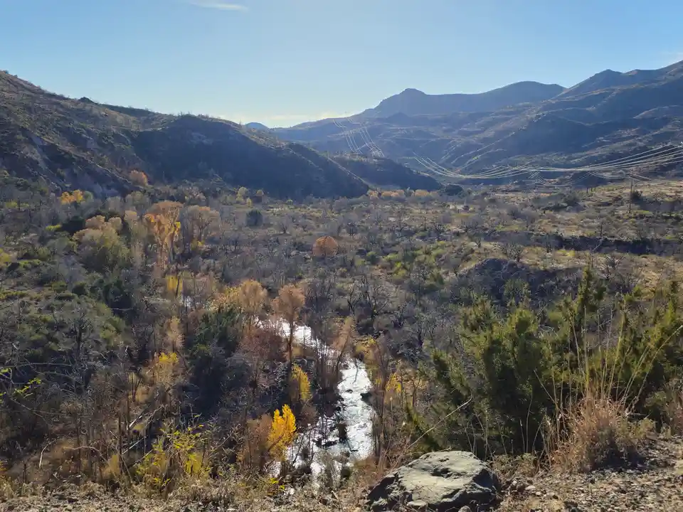 A scenic photo of a river flowing through a dry, hilly valley with autumn-colored trees and power lines stretching across…