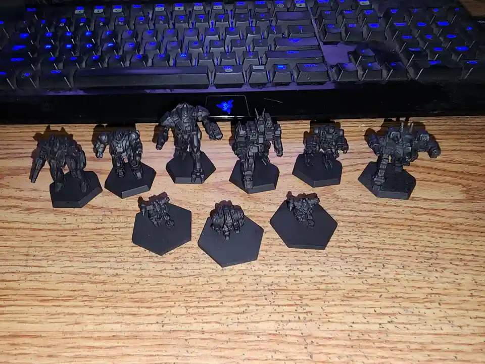 A photo shows eight large, detailed, grey robot miniatures standing on hexagonal bases in a row on a wooden desk, with a…