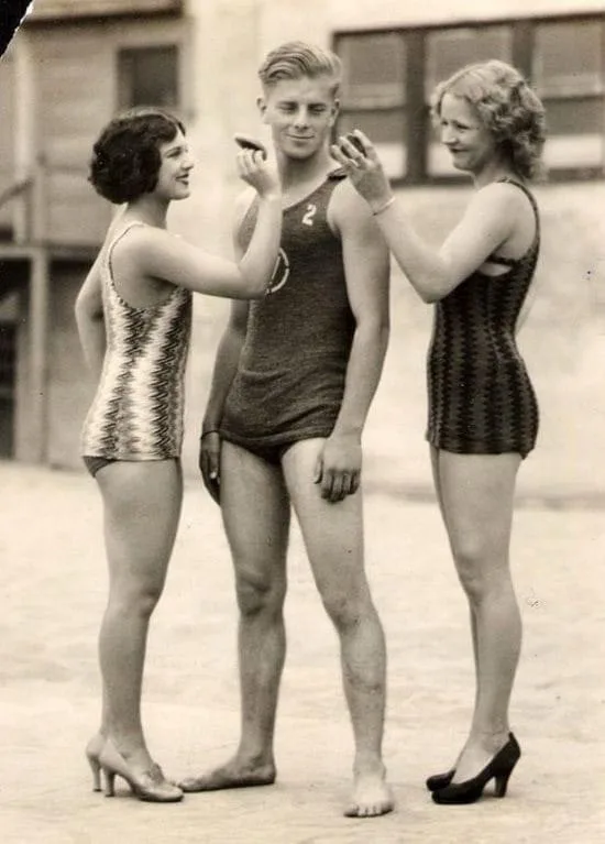 A vintage black-and-white photo shows two women in patterned swimsuits applying makeup to a man's face while he smiles; one…