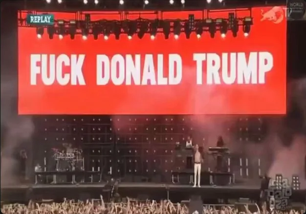 A large red screen at a concert displays the text 'FUCK DONALD TRUMP' in bold white letters. Below, a performer stands on…
