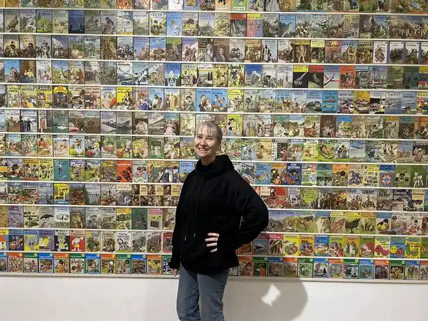 A woman in a black hoodie and jeans stands smiling with one hand on her hip in front of a massive wall covered in colorful…