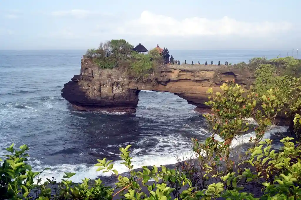 A scenic photo of the Tanah Lot temple in Bali, perched on a rocky island with waves crashing below and lush greenery in the…