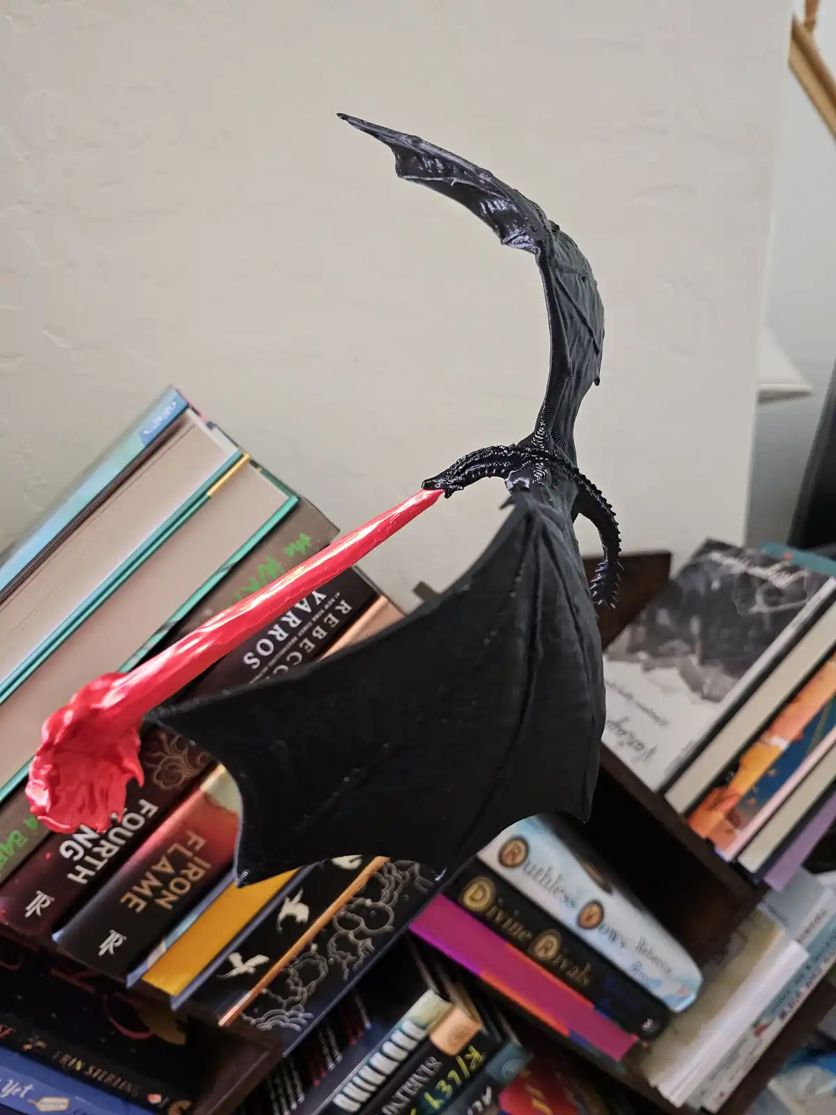 A small black dragon figurine with spread wings is perched on a stack of books, its mouth open to reveal a bright red…